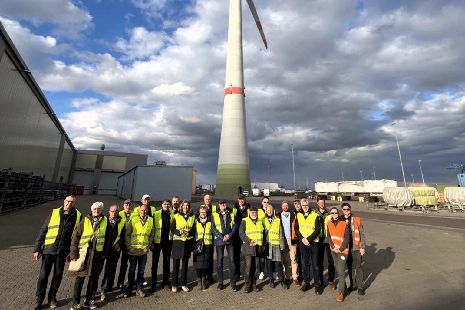 Personengruppe mit Warnwesten vor Windkraftanlage und Industriegebäuden bei bewölktem Himmel.