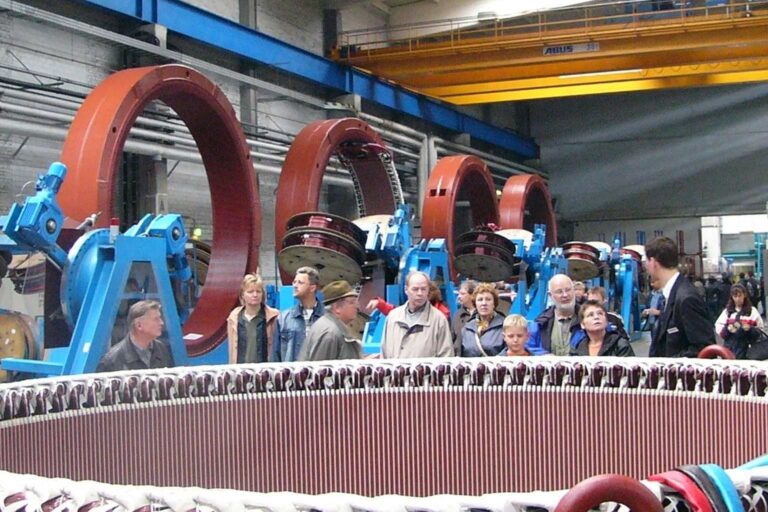 Gruppe von Menschen besichtigt große rote Maschinenringe in einer Fabrikhalle.