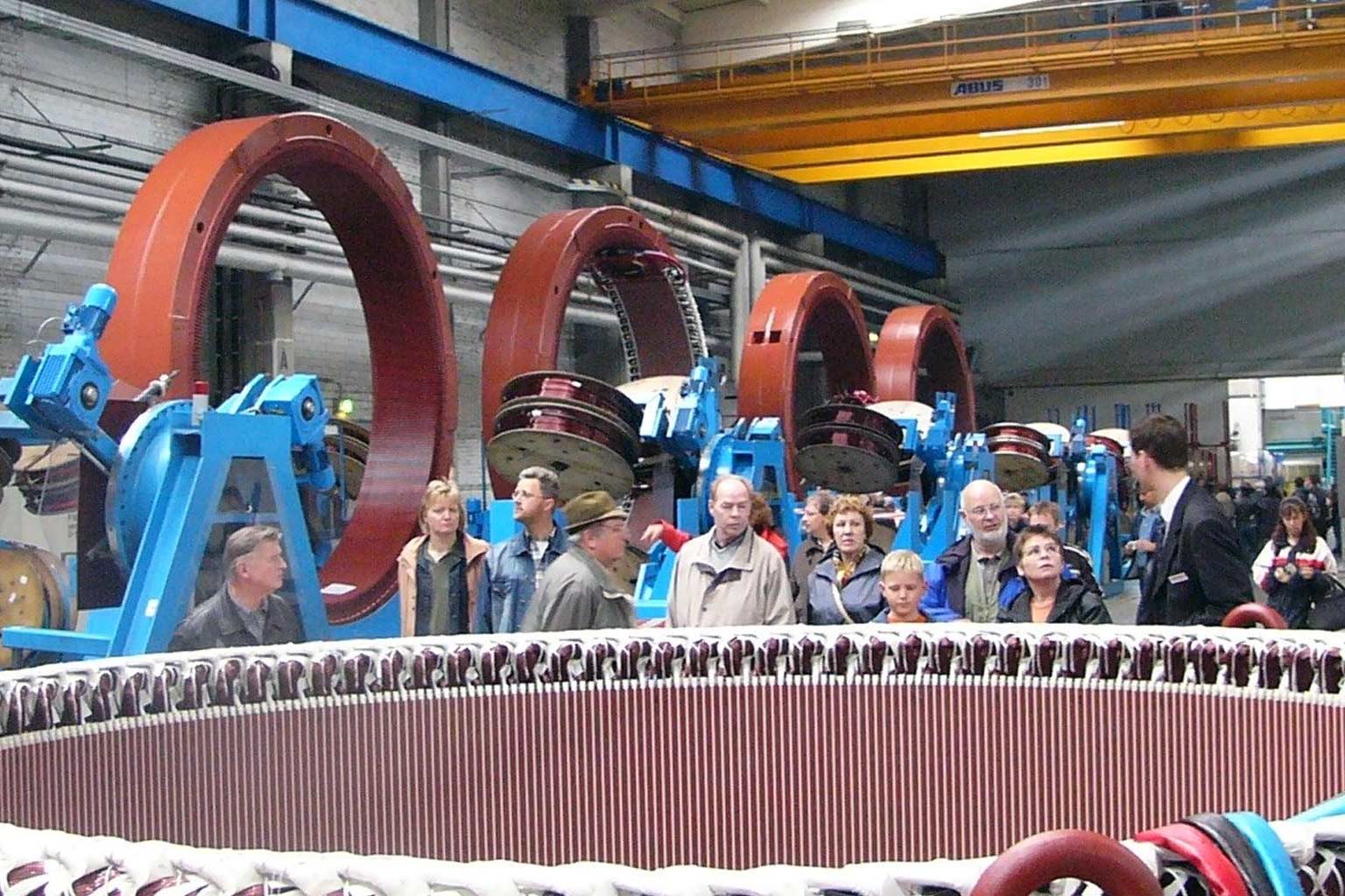 Gruppe von Menschen besichtigt große rote Maschinenringe in einer Fabrikhalle.