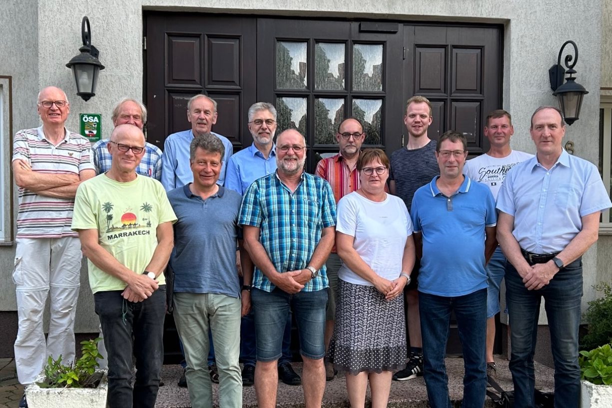Gruppe von 13 Personen steht vor einer Eingangstür, zwei Außenlampen flankieren die Szene.