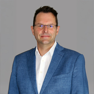 Mann in blauem Anzug und weißem Hemd vor grauem Hintergrund.