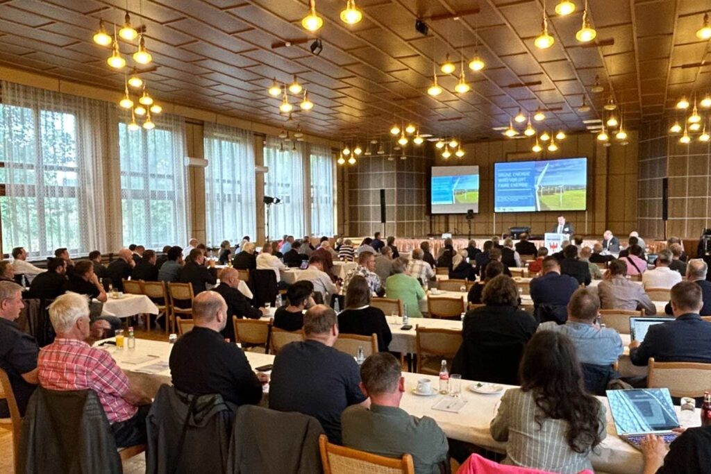 Großer Konferenzraum mit vielen Menschen an gedeckten Tischen, Vortrag auf zwei Bildschirmen vorne.