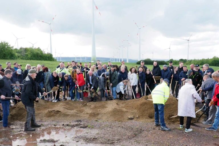 Eine Gruppe von Menschen beim Spatenstich auf einer Wiese mit mehreren Windrädern im Hintergrund.