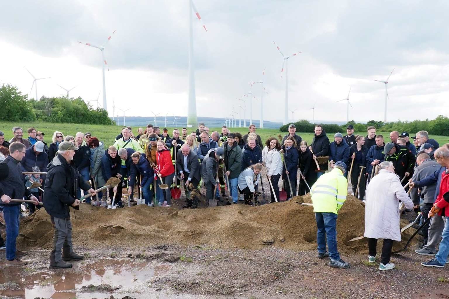 Eine Gruppe von Menschen beim Spatenstich auf einer Wiese mit mehreren Windrädern im Hintergrund.